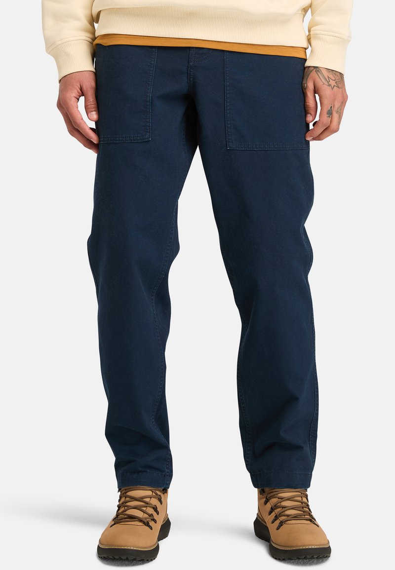 Navy blue, relaxed-fit cargo pants made of durable cotton with two front pockets; paired with tan hiking boots, featuring lace-up design.