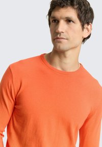 Fel oranje gebreide trui, ronde hals, aansluitend ontwerp, zachte textuur, met zichtbare naden op de schouders en subtiele geribbelde details bij de halslijn.