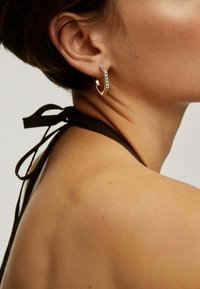 Mujer con un pendiente de aro hexagonal dorado con texto grabado, blusa sin mangas marrón anudada al cuello, mostrada desde un ángulo cercano lateral.