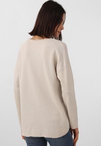 Pull beige à coupe décontractée, doté d'un col rond et de détails à ourlet effiloché. Les manches longues et le tissu doux et texturé complètent le design.