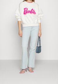 Vit sweatshirt med rosa "Barbie"-logotyp, ljusblå utsvängda jeans, lavendelfärgade klacksandaler och en liten denimhandväska.