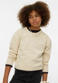 Suéter de punto color crema con cuello en V, borde negro acanalado y mangas tres cuartos, combinado con una falda negra a rayas y un collar de cuentas.