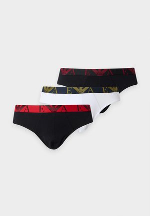 Homme aux cheveux foncés, debout pieds nus, portant un slip noir avec une ceinture orange sur un fond blanc uni.