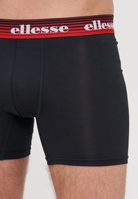 Boxer a vita bassa neri con un design aderente, dotati di una fascia elasticizzata rossa e bianca a righe con il logo 'ellesse'. Texture liscia.