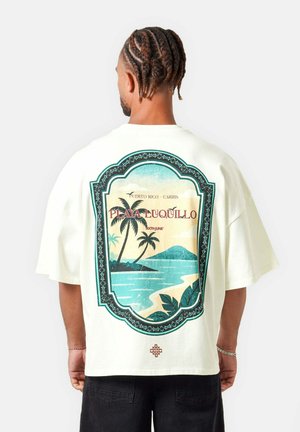Man met ingevlochten haar die een wit T-shirt draagt met een tropische strandprint en de tekst "Playa Luquillo, Puerto Rico - Caribs" op de achterkant.