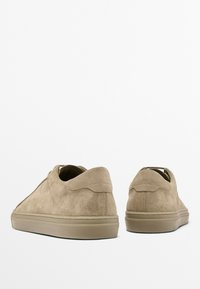 Beige suede sneakers with rounded toe, low profile, minimal seams, and a textured finish. Flat rubber sole and lace-up design.