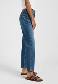 Jeans in denim blu con taglio a gamba dritta, dotati di cinque tasche e patch con logo sul retro. Abbinati a sandali scivolati rossi, con dettagli delle cinghie texture.