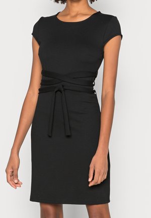 Woman wearing a short-sleeve black dress with a round neckline and a wrap-around belt tied at the waist.