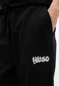 Schwarze Baumwoll-Jogger mit elastischem Bund und Kordelzug. Mit weißem gesticktem "Hugo"-Logo auf der linken Seite. Glatte Textur.
