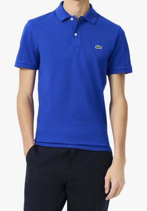 Homme portant un polo bleu vif à manches courtes avec un petit logo crocodile vert sur la poitrine gauche, associé à un pantalon noir.