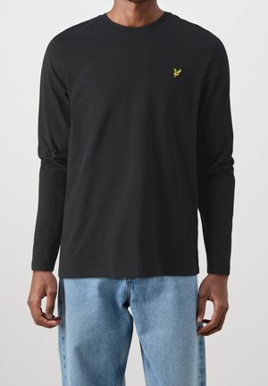 Mann trägt schwarzes Langarmshirt mit kleinem gelbem Vogel-Logo auf der Brust und hellblauen Jeans, steht vor neutralem Hintergrund.