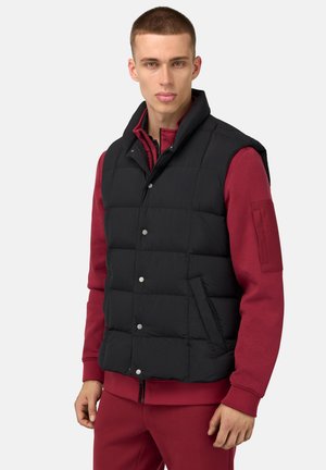 Schwarze Pufferweste mit Druckknöpfen, hohem Kragen und vorderen Taschen, getragen über einem roten Langarmshirt. Glatte, gesteppte Textur.