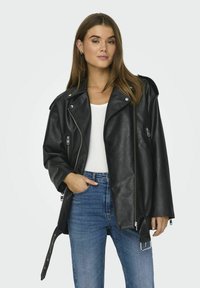Zwarte leren motorjack met zilveren ritsen, brede revers en een oversized pasvorm. Gecombineerd met een witte top en blauwe jeans.