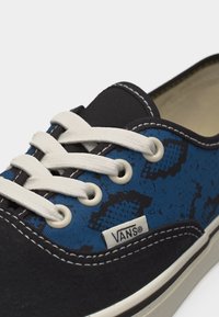 Baskets Vans bleu et noir avec des lacets blancs, des œillets blancs, des coutures visibles et une petite étiquette du logo Vans sur le côté.