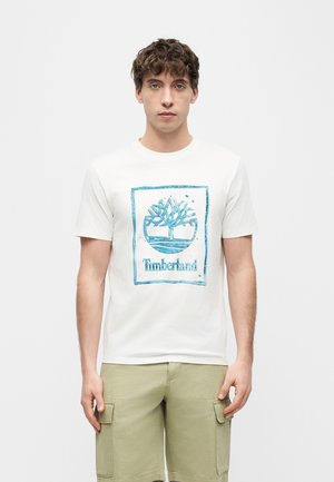Jeune homme portant un t-shirt blanc avec le logo Timberland et un short cargo beige, debout devant un fond blanc uni.