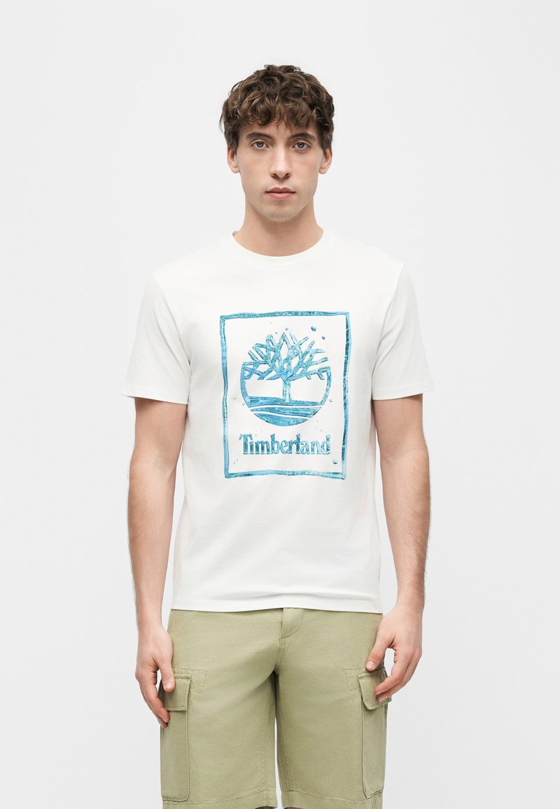 Νεαρός άνδρας που φοράει λευκό T-shirt με το λογότυπο Timberland και μπεζ σορτς cargo, στέκεται μπροστά σε απλό λευκό φόντο.