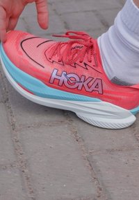 Hoka futócipő élénk korall színű felsőrésszel, fekete akcentussal és világoskék talppal. A texturált hálós anyag, vastag párnázás és kiemelkedő logó jellemzi.