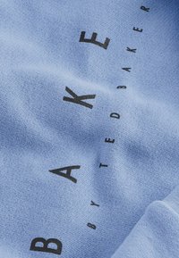 Světle modrá látka má vyražený text "BAKE BY TED BAKER" v černé barvě. Textura vypadá jemně a mírně texturovaně.