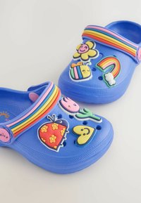 Blaue Clogs mit einem Regenbogenriemen, 3D-farbigen Verzierungen, einschließlich Blumen, Herzen und einer Biene, mit Belüftungslöchern und strukturierten Sohlen.