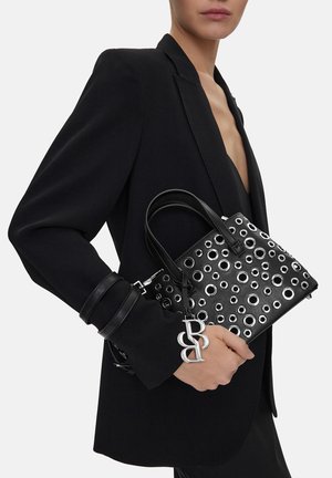 Mujer con blazer negro sosteniendo un bolso de cuero negro con ojales plateados y un adorno metálico de la marca, sobre fondo blanco.