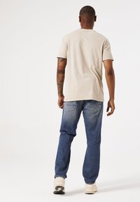 Beige Baumwoll-T-Shirt mit Rundhalsausschnitt, kombiniert mit blauen Jeans. Die Jeans haben eine ausgewaschene Optik und Gesäßtaschen. Cremefarbene Sneakers runden den Look ab.