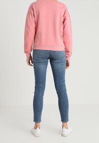 Persona che indossa un felpa rosa, jeans skinny blu e sneaker bianche, in piedi contro uno sfondo chiaro e uniforme, girato di spalle.