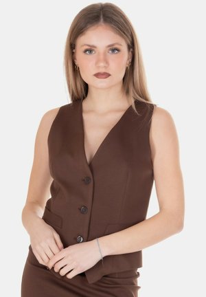 Giovane donna con capelli biondi lisci indossa un gilet marrone senza maniche con bottoni e gonna abbinata, in piedi davanti a uno sfondo bianco.