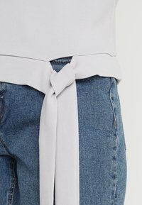 Lichtgrijze top met een knoopdetail bij de taille, gecombineerd met blauwe denim jeans. De top heeft een zachte textuur en een subtiele glans.