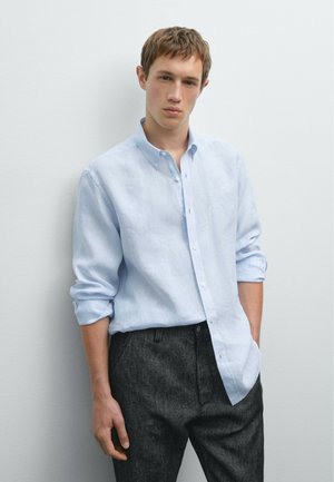 Light blue button-up shirt with long sleeves and a relaxed fit, paired with dark gray trousers, resting against a neutral background.