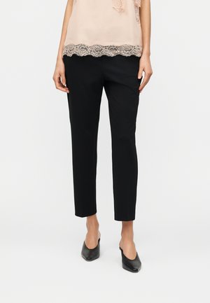 Žena stojící na bílém pozadí, oblečená v černých úzkých kotníkových kalhotách, bledě růžovém topu s krajkovým lemem a černých botách s špičatou špičkou a nazouvacím stylem.