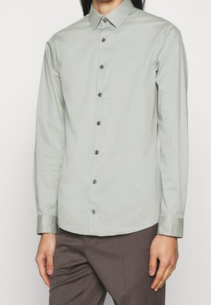 Homme portant une chemise gris clair à manches longues avec col et un pantalon gris foncé, debout devant un fond uni.