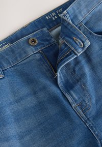 Jean en denim bleu coupe slim avec fermeture éclair standard, bouton en métal, et détails de couture visibles. L'étiquette indique la taille 34.