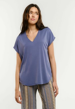 Young woman with long brown hair wearing a loose blue V-neck top and patterned wide-leg pants, looking slightly to the side.