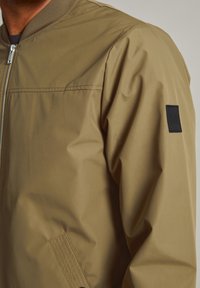 Chaqueta bomber color caqui con una textura suave y ligera, cierre de cremallera, cuello de canalé y una pequeña etiqueta negra en la manga izquierda.