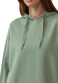 Hellgrüner Hoodie mit lockerer Passform, ausgestattet mit einer Kordelzugkapuze und strukturiertem Stoff. Auffällige Details sind die gebrandeten Kordeln.