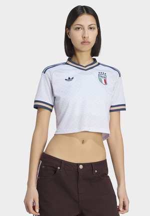 Mladá žena s rovnými tmavými vlasy má na sobě krátký bílý italský fotbalový dres a tmavé kalhoty, stojí před jednoduchým světlým pozadím.