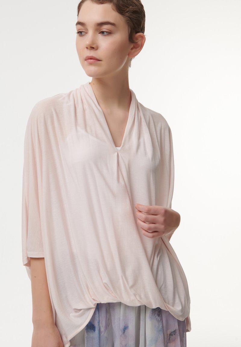 Lichtroze, losvallend top met een gedrapeerd ontwerp en V-hals. Zachte textuur, korte mouwen en geraffineerde details aan de zoom.