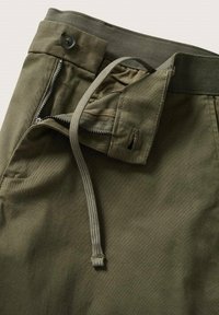 Gros plan sur un pantalon vert olive avec une taille boutonnée, une braguette zippée et un détail de cordon sur un fond uni.