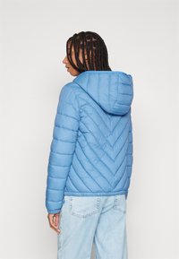 Světle modrá prošívaná bunda s chevronovým prošíváním, kapucí a elastickými manžetami, nošená s světle modrými džínami. Hladká textura a lehký design.