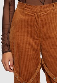 Bruine corduroy broek met verticale ribbels, voorzien van drukknopen aan de zijkant. Het materiaal lijkt zacht met een gestructureerde afwerking.