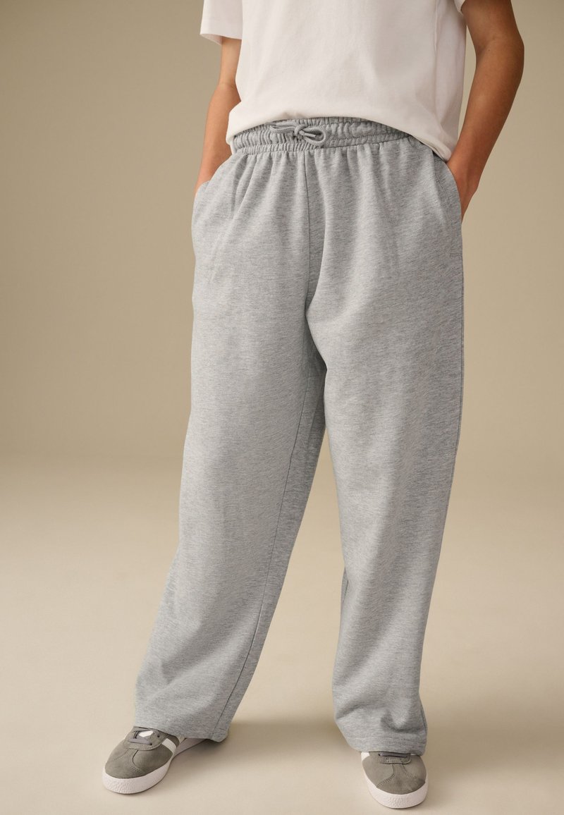 Personne portant un pantalon de survêtement ample gris clair avec une taille élastique à cordon et des baskets grises, debout les mains dans les poches devant un fond uni.