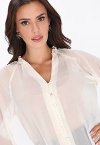 Blusa ligera y transparente color crema con un escote con volantes y cierre frontal de botones. Presenta una textura suave y mangas largas con plisados delicados.