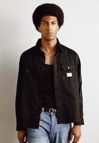 Chemise noire à boutons avec deux poches poitrine, portée sur un débardeur noir. Associée à un jean bleu clair, affichant un style décontracté.