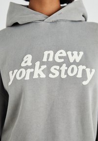 Szürke kapucnis pulóver, elülső zsebbekkel, húzózsinórral ellátott kapucnival és fehér textúrált betűzéssel, amely "a new york story" feliratot tartalmaz. Puha szövettextúra.