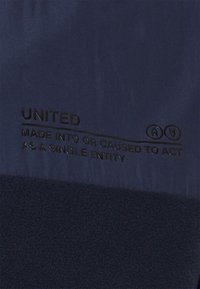 Marineblauer Stoff mit einem strukturierten Fleece-Bereich. Schwarzer gedruckter Text lautet „UNITED MADE INTO OR CAUSED TO ACT AS A SINGLE ENTITY.“