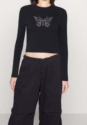 Femme portant un top court noir à manches longues avec un motif papillon en strass et un pantalon noir ample sur un fond uni.
