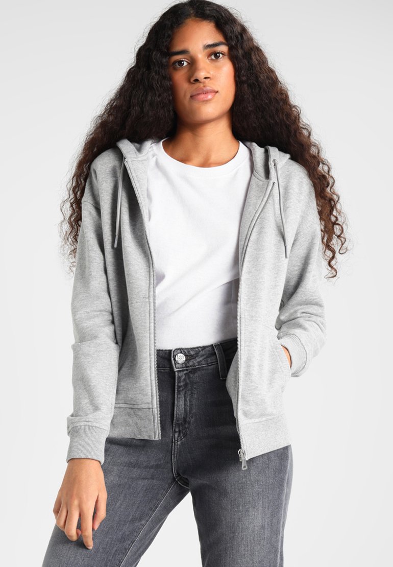 Joven mujer con cabello largo y rizado, vestida con una sudadera con capucha gris con cremallera, camiseta blanca y vaqueros gris oscuro sobre un fondo liso.
