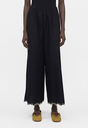 Personne portant un pantalon noir large avec des ourlets festonnés et des détails œillets, associé à des ballerines Mary Jane tissées jaune moutarde.