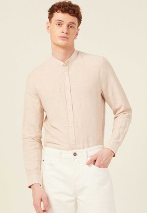 Camicia a maniche lunghe beige chiaro con colletto alla cinese e bottoni frontali, indossata con pantaloni bianchi. Il tessuto appare morbido con una leggera texture.