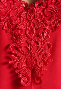 Blusa vermelha com intrincados bordados florais na gola, com um tecido suave e texturizado e um padrão de renda detalhado.